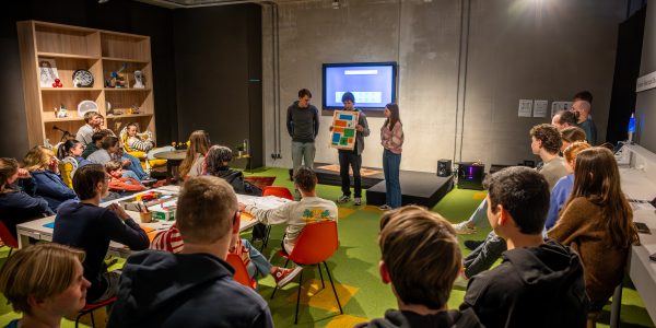 Groep scholieren in een ruimte die kijken naar drie scholieren die iets presenteren.